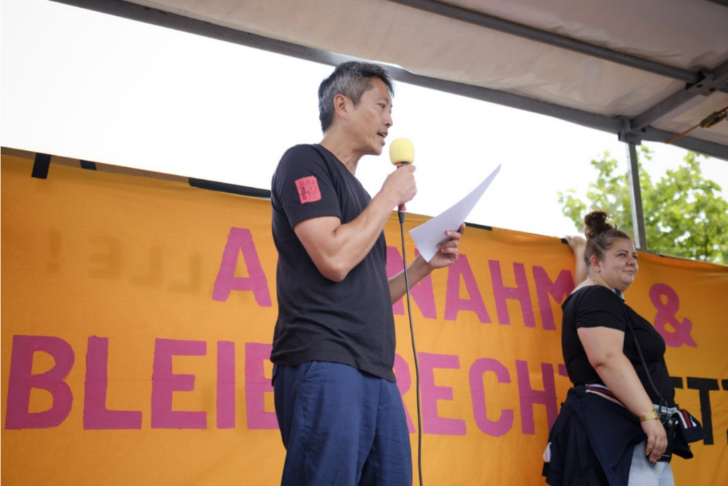 picture: 🔗 Kien Nghi Ha speaking at the national demonstration marking the 30th anniversary of the pogrom in Rostock-Lichtenhagen on 27 August 2022. photo: Dong-Ha Choe.
